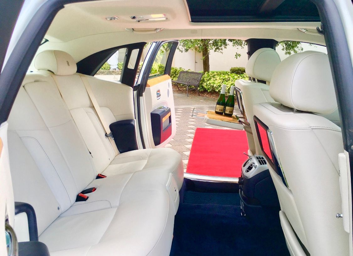 The interior of a luxury car with white leather seats, dark carpet, and a red rug on the ground outside the open door.