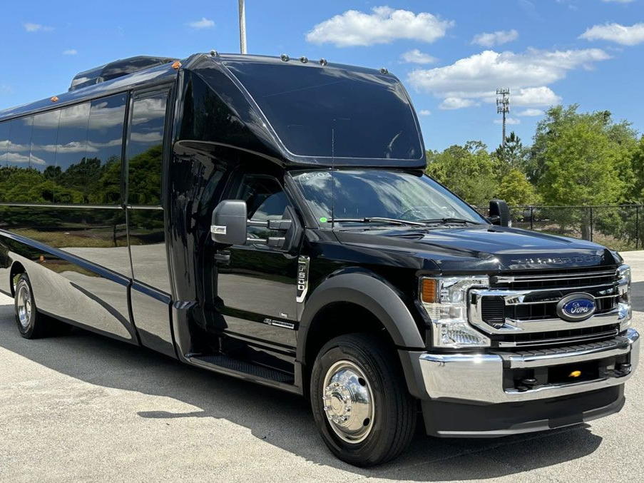 A black Ford luxury party bus parked outdoors on a sunny day with blue sky and trees in the background.