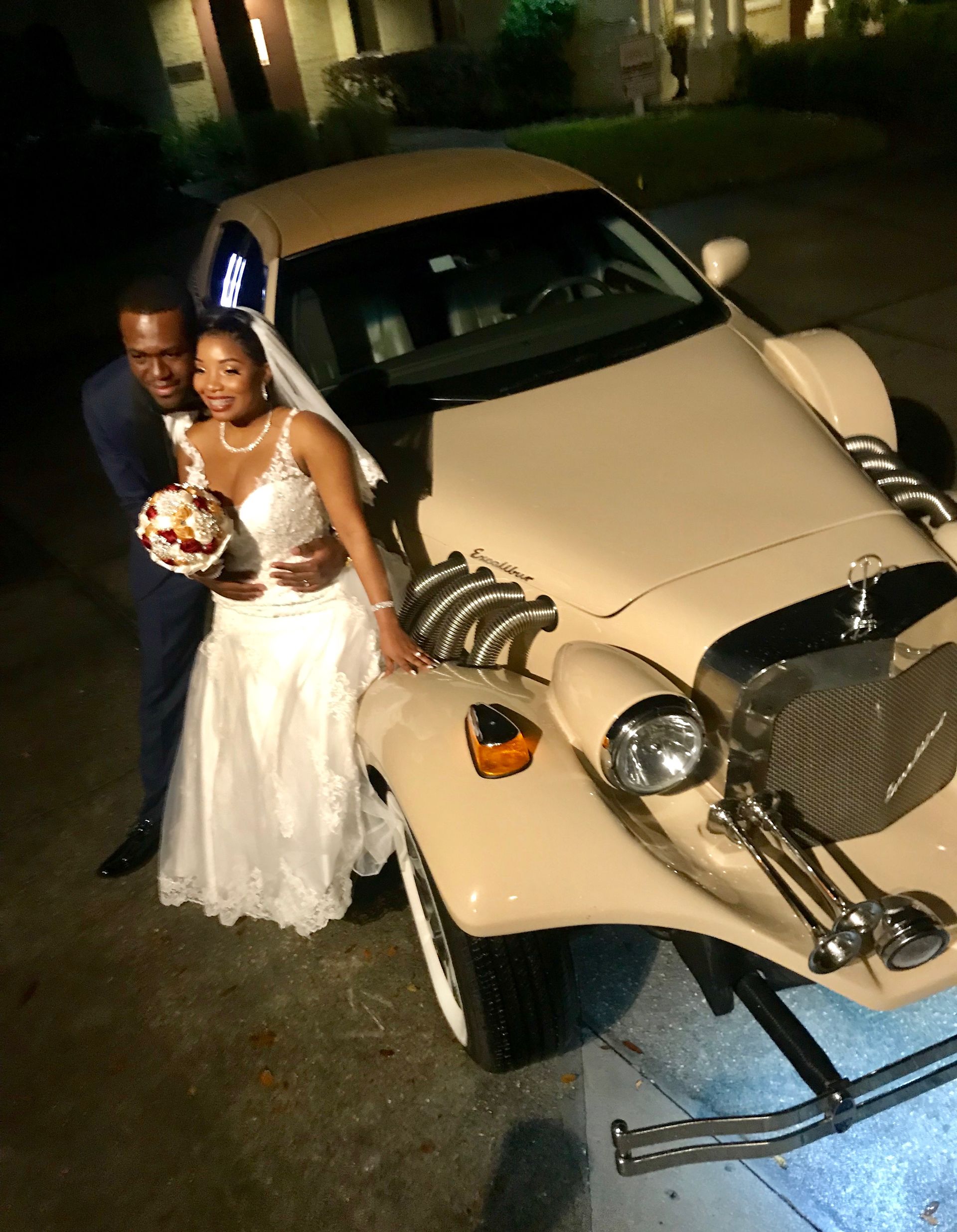 A couple in wedding attire poses next to a vintage, tan, convertible car at night.