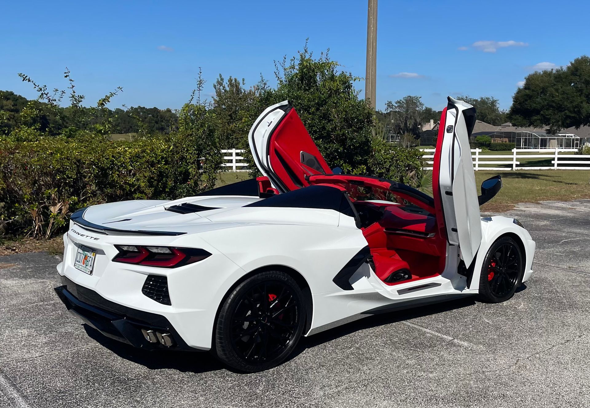 A white Chevrolet Corvette with red interior and vertical doors parked on a gravel lot under a clear blue sky.
