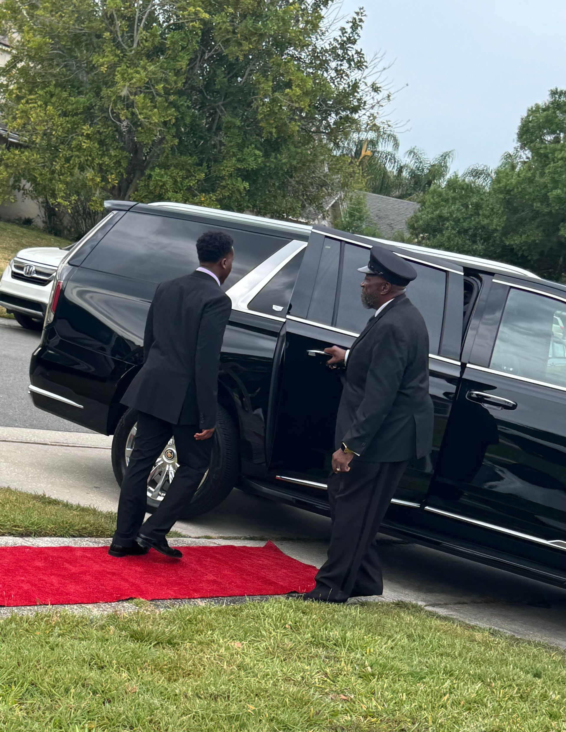 A chauffeur in uniform opens the rear door of a black SUV parked by a red carpet on a lawn.
