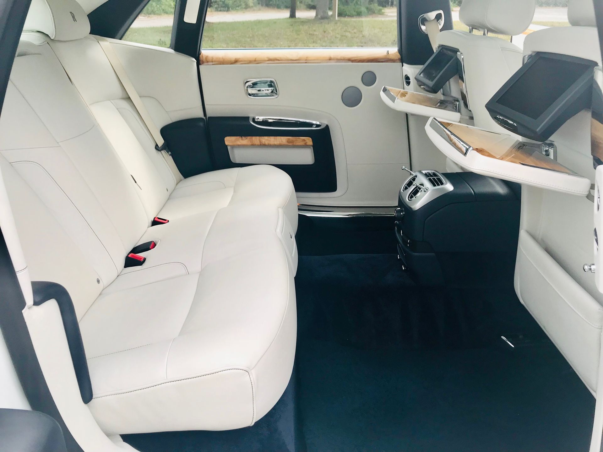 Interior of a luxury car with white leather seats, dark floor mats, wooden trim, and seat-back mounted media screens.