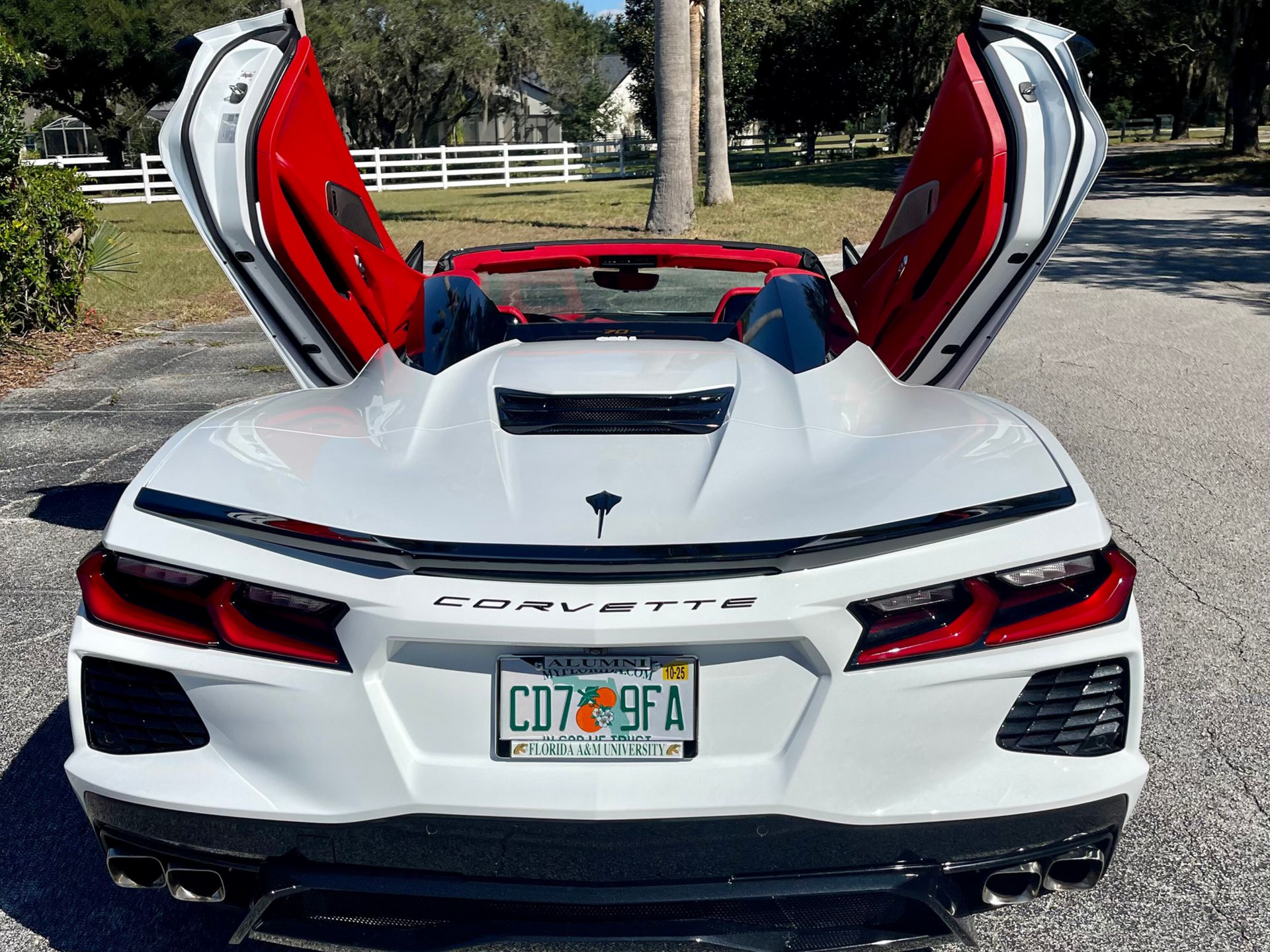 A white Chevrolet Corvette convertible with its red-lined gullwing doors open, parked on a paved driveway.
