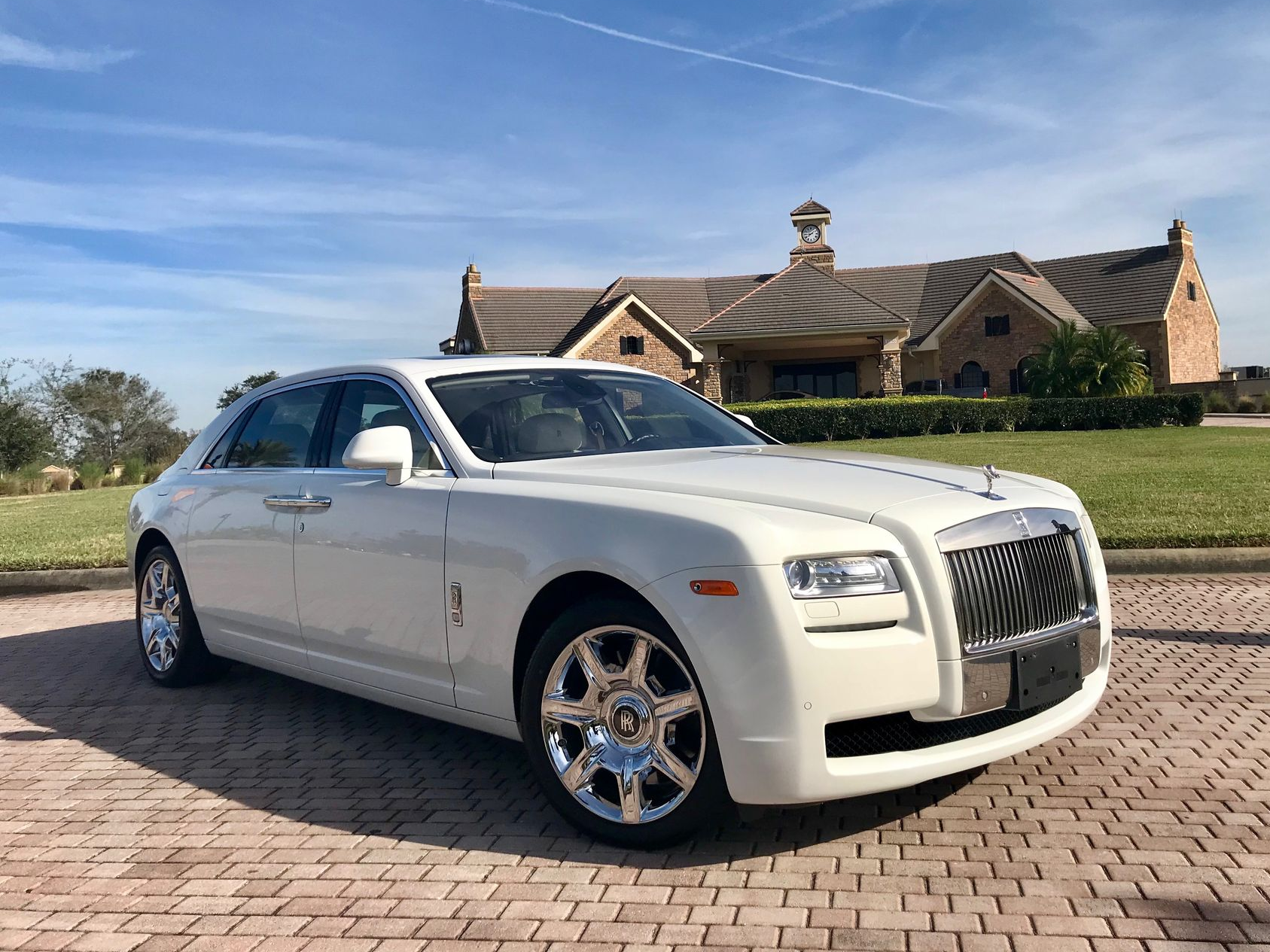 A white Rolls-Royce sedan parked on a brick driveway in front of a stone mansion under a sunny blue sky.