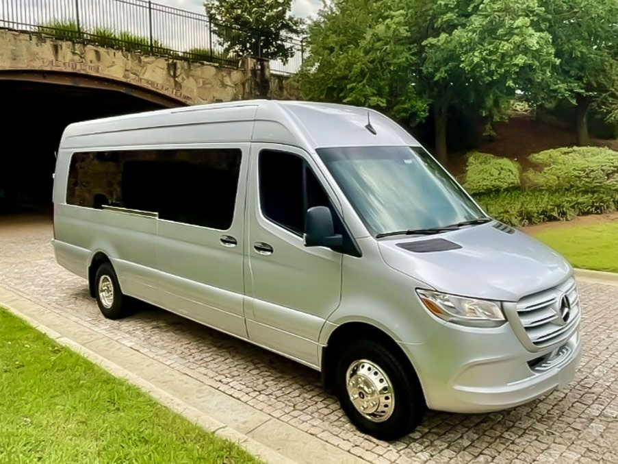 Silver Mercedes-Benz Sprinter passenger van parked on a cobblestone path near a stone bridge and lush green trees.