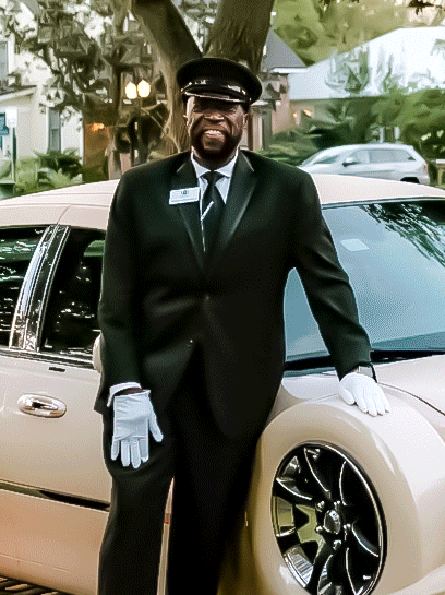 A chauffeur in a black suit, white gloves, and cap stands smiling beside a cream-colored vintage car on a paved street.