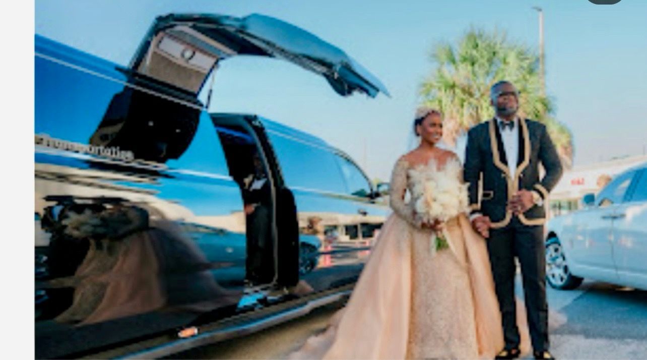 Bride and groom pose by a black limousine with open doors on a sunny day.