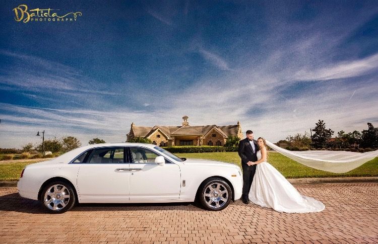 Bride and groom pose by a white Rolls Royce on a brick road; building in the background.