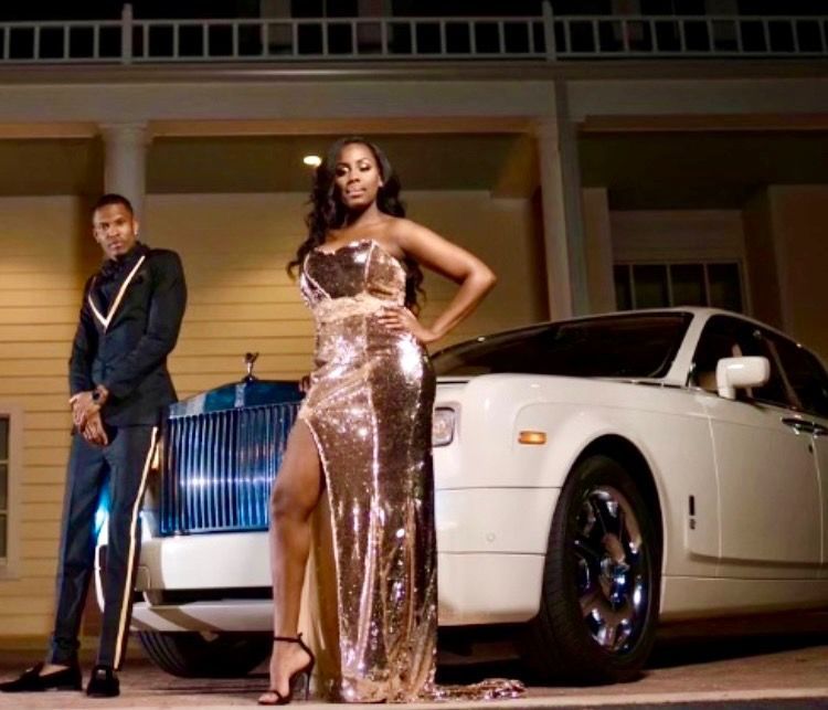 A person in a shimmering gold gown poses in front of a white Rolls-Royce with a person in a dark suit standing nearby.