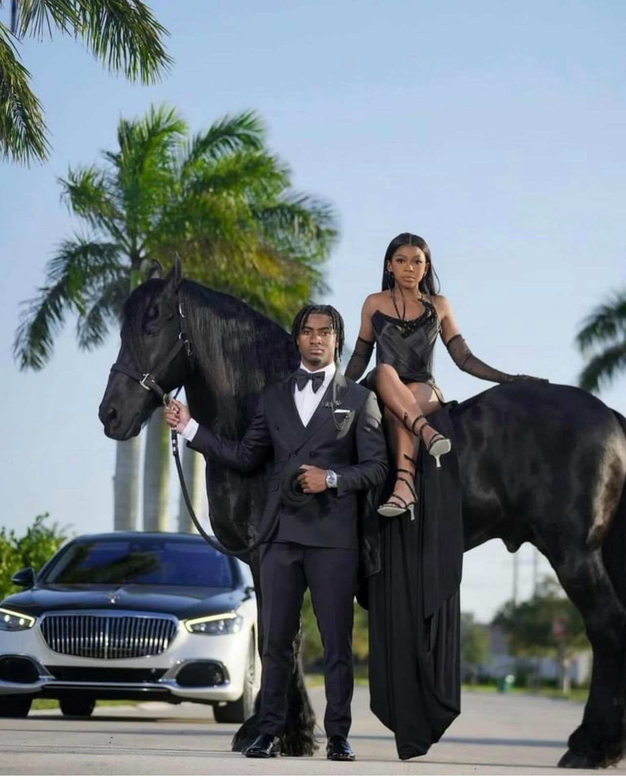 A person in a suit stands next to a black horse carrying a person in a formal black dress, with a car and palms behind.