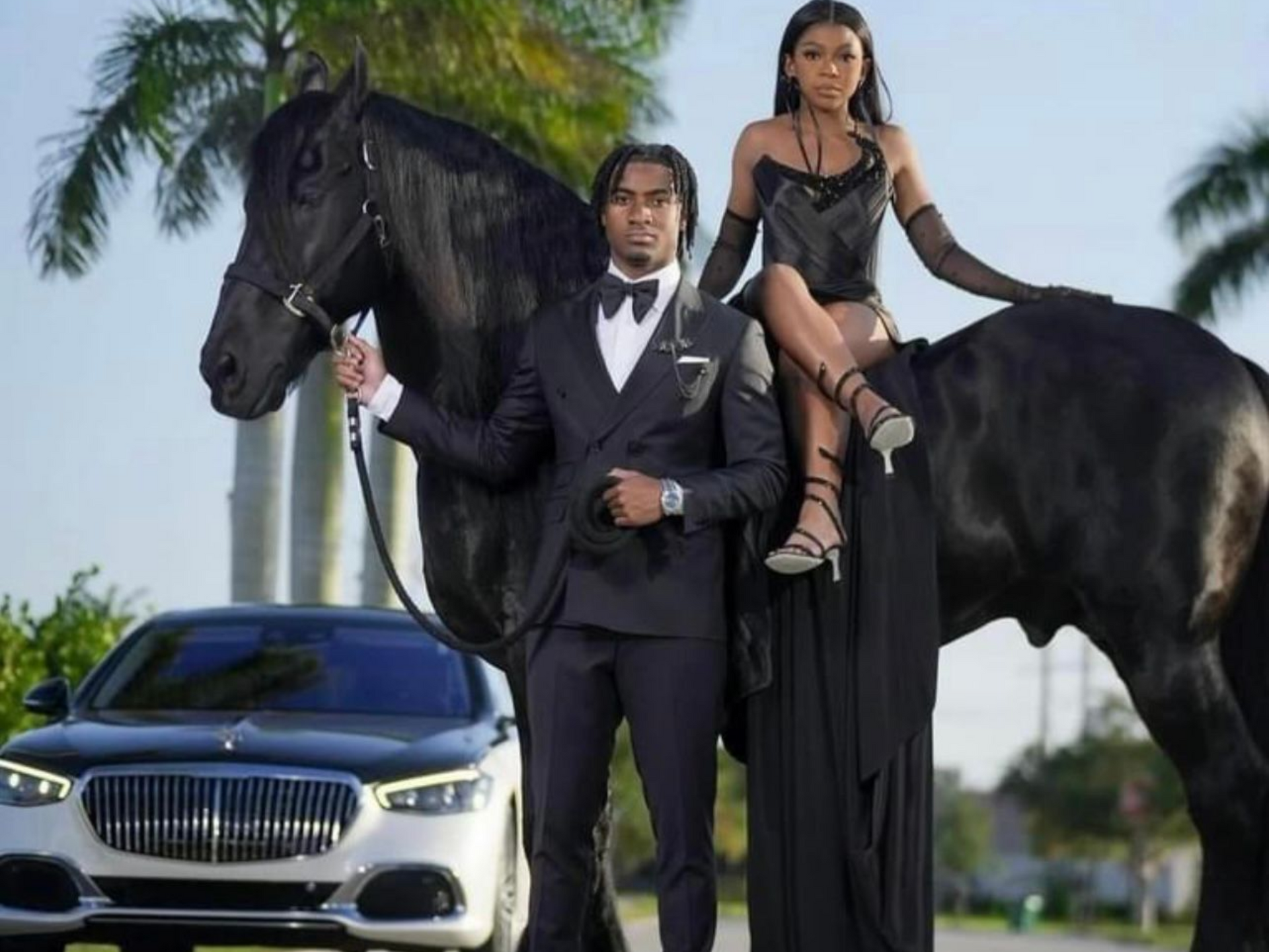 A person in a suit stands beside a horse as another person in a black gown sits on the horse, with a car parked nearby.