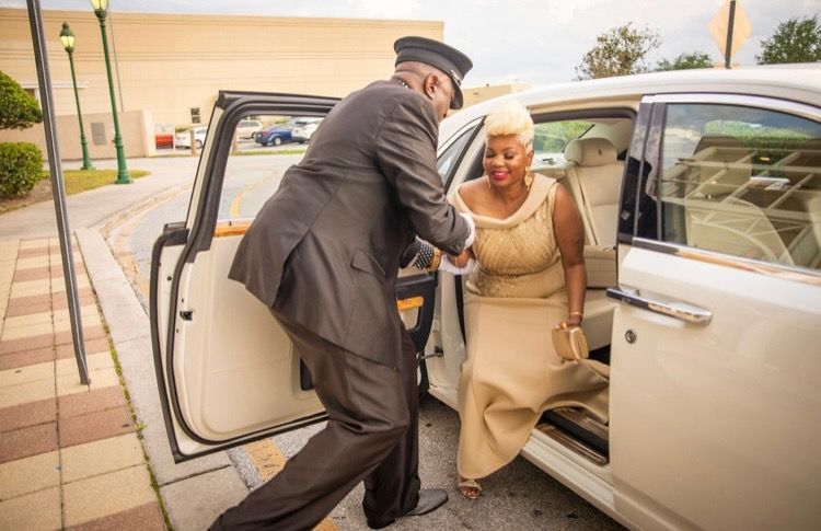 A chauffeur in uniform assists a person in a formal gold dress as they step out of a white luxury car.