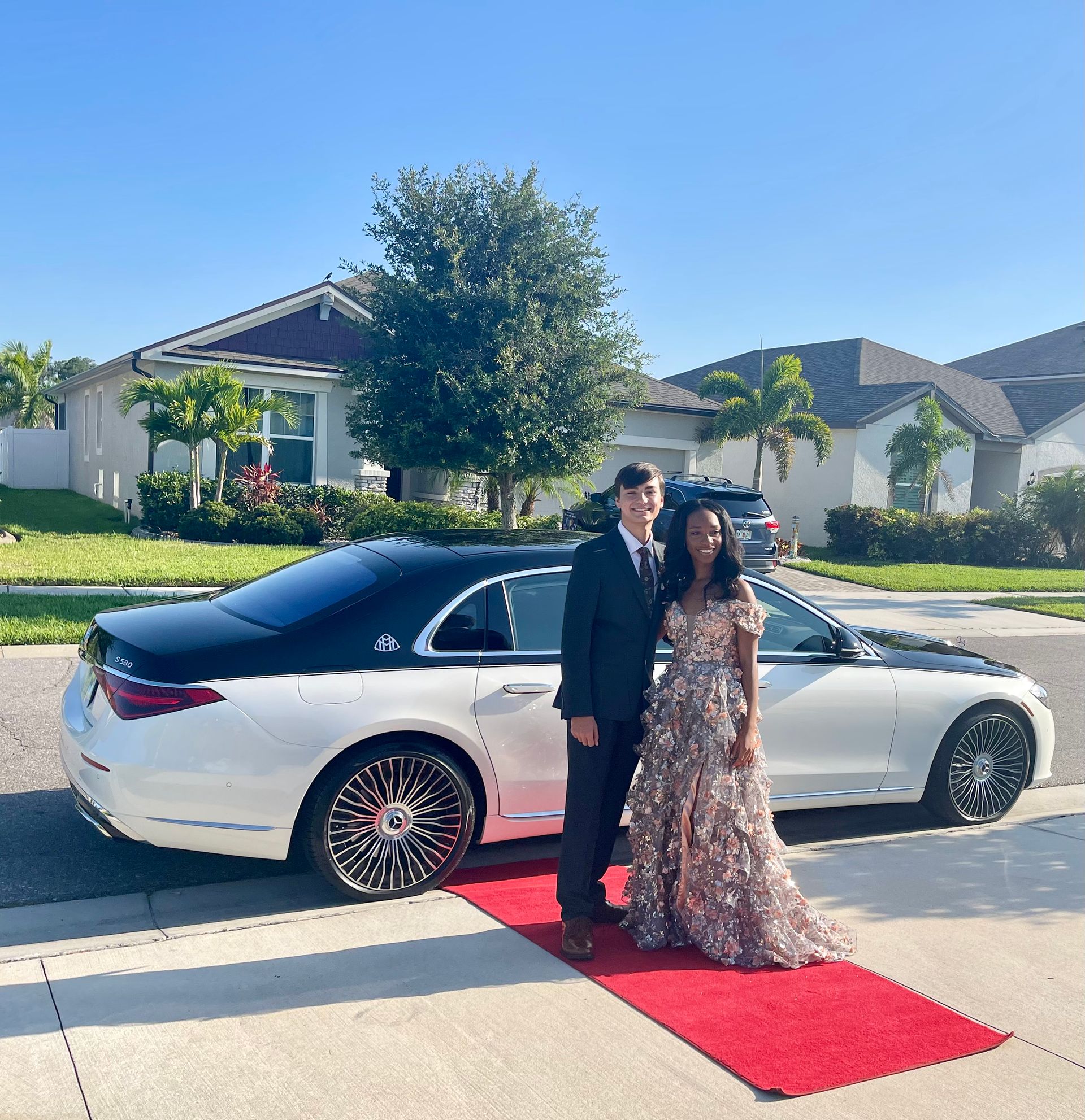 A formal couple poses on a red carpet next to a two-tone white and black luxury sedan in a residential suburban setting.