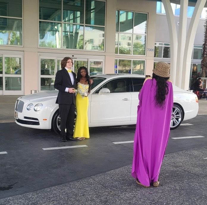 A person in a magenta dress photographs a couple in formal wear next to a white luxury car outside a modern building.