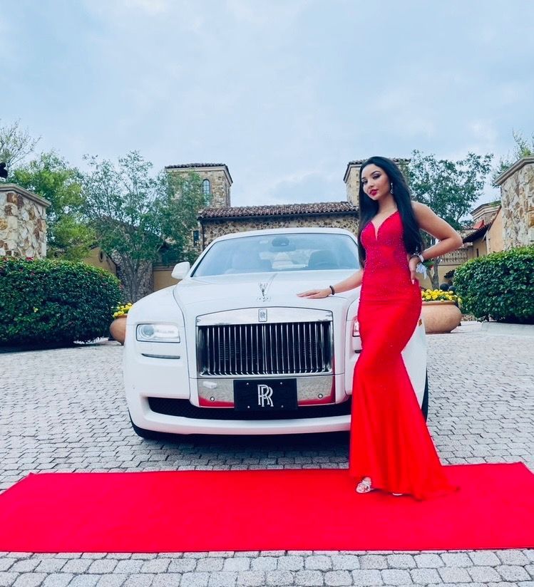 A person in a long red dress poses on a red carpet next to a white Rolls-Royce in a stone-paved driveway.