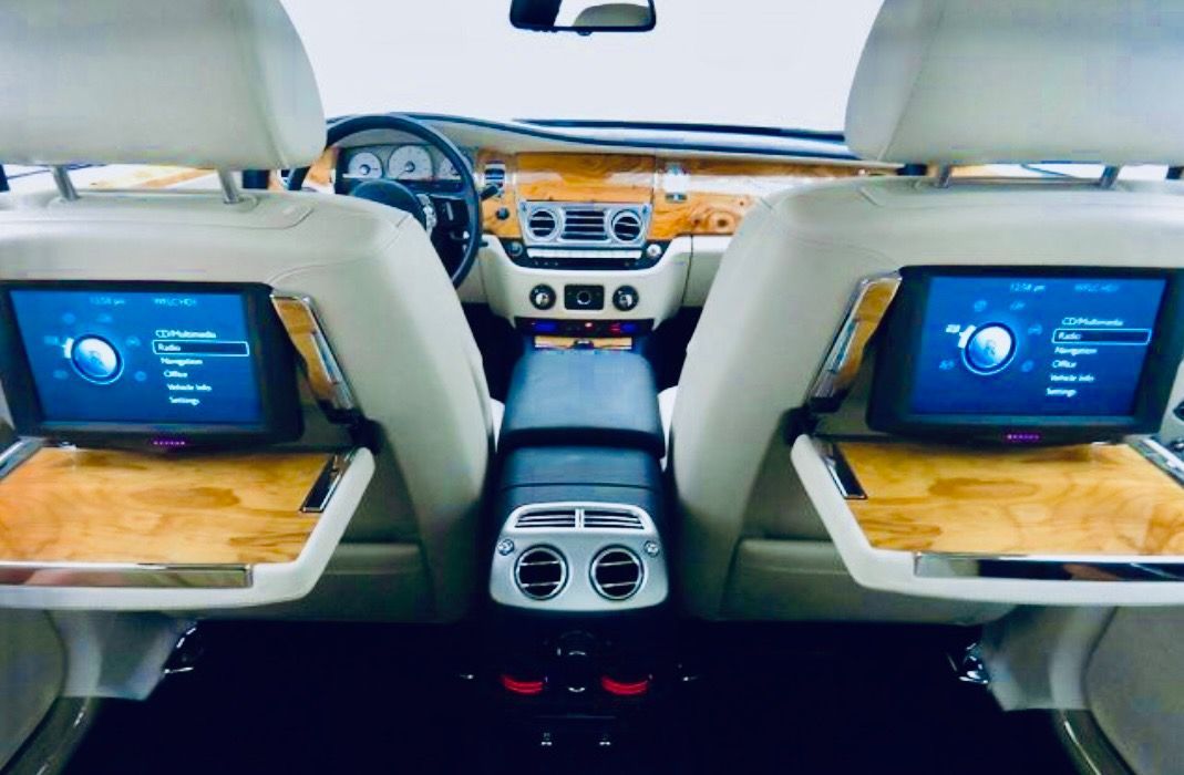The back seat of a luxury car interior features dual mounted screens and wood veneer folding tray tables.