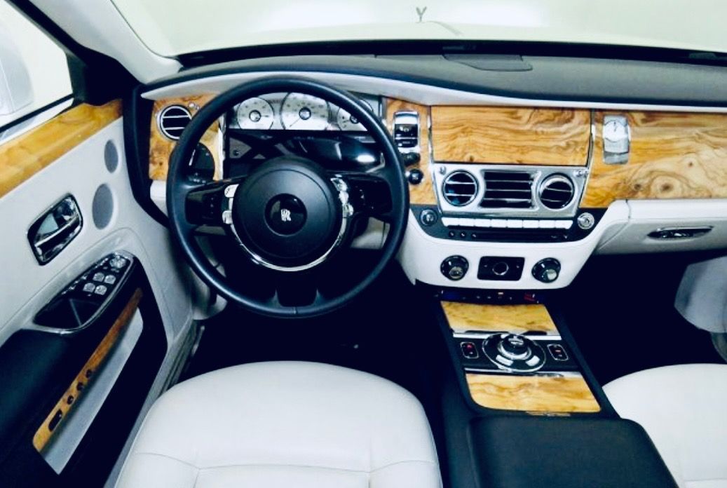 The interior of a Rolls-Royce, featuring a black steering wheel, white leather seats, and light wood dashboard trim.
