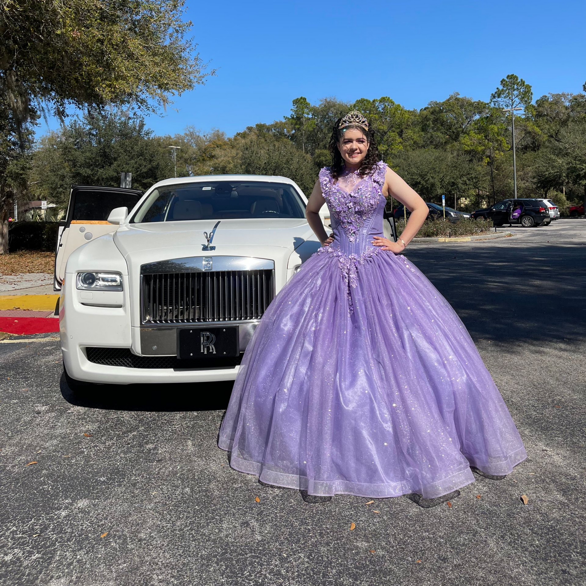 A person in a glittery purple ball gown and tiara poses standing next to a white Rolls-Royce in a parking lot.