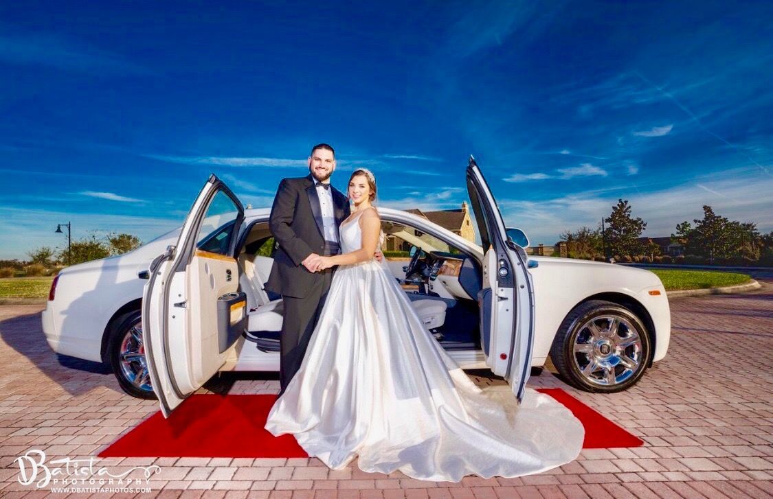 A bride and groom pose by a white car with open doors on a red carpet outdoors under a vibrant blue sky.