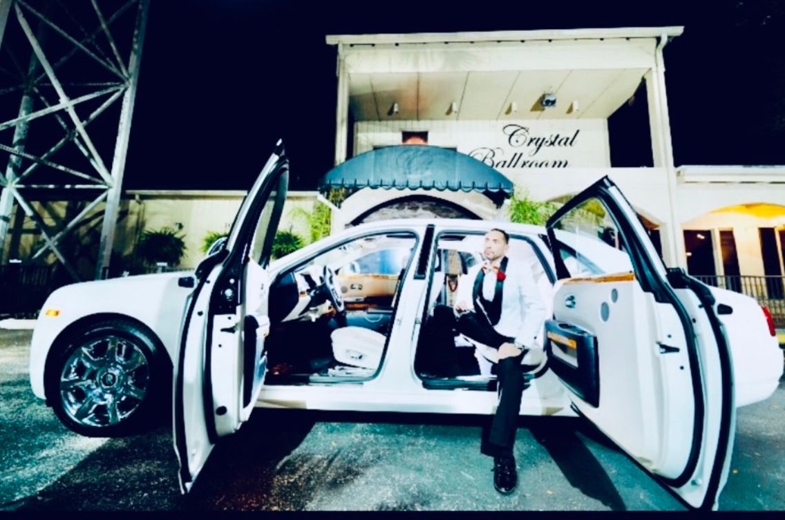 A person in a formal white suit sits in the open rear door of a white luxury car at night in front of the Crystal Ballroom.
