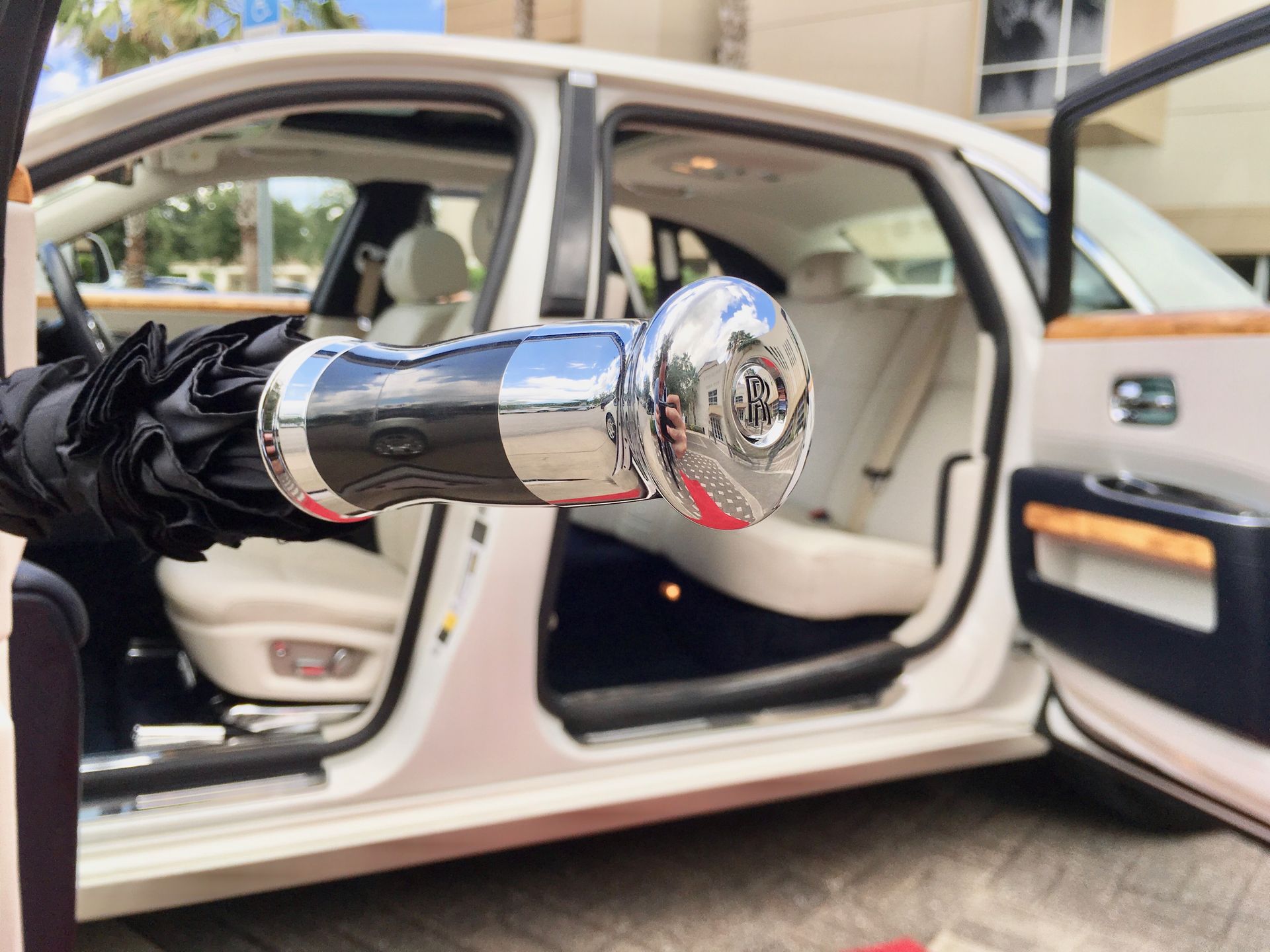 A hand holds a luxury Rolls-Royce umbrella handle with a silver emblem, with a white sedan door open in the background.