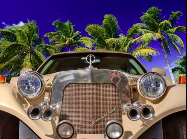 Close-up front view of a beige vintage Excalibur car with chrome accents parked in front of tropical palm trees.