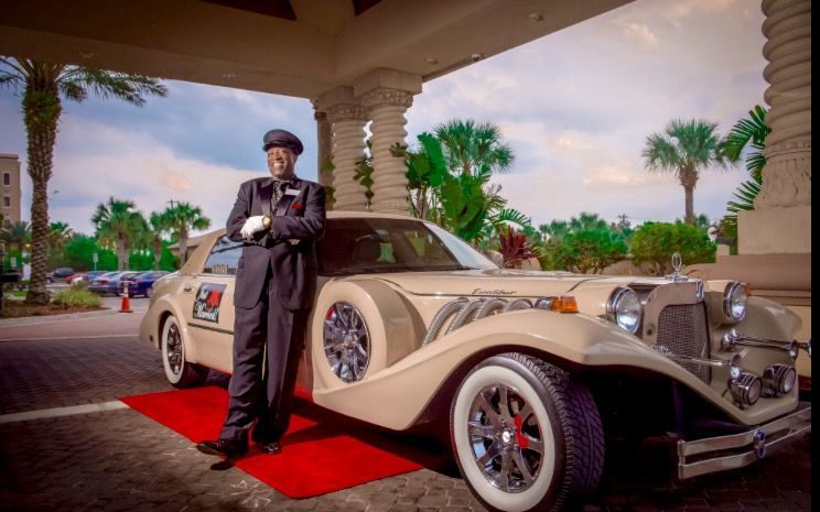 A valet in a uniform leans against a vintage tan luxury car parked on a red carpet under a building entrance.