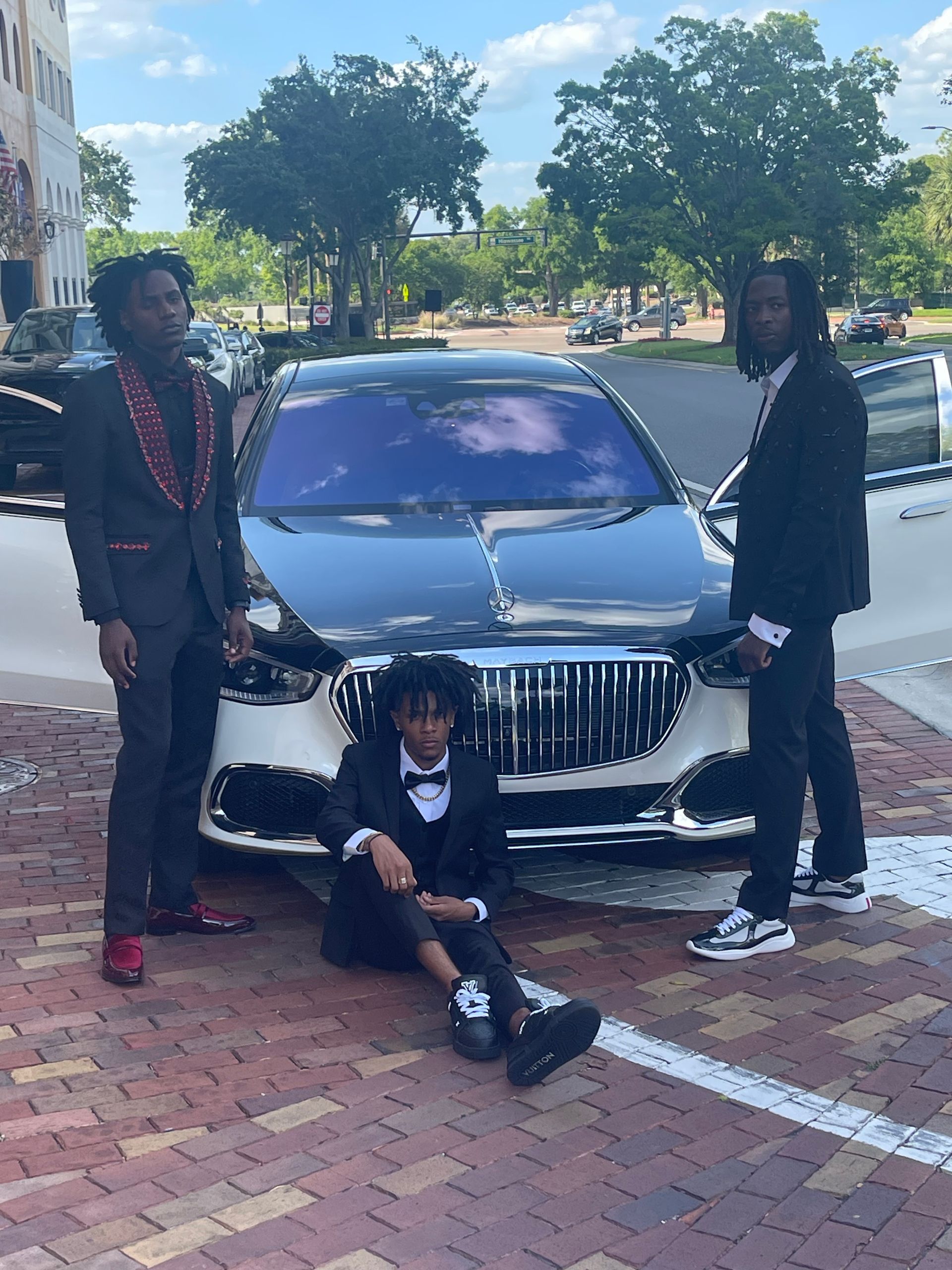 Three people in formal attire pose around a luxury black and white car on a brick-paved surface.