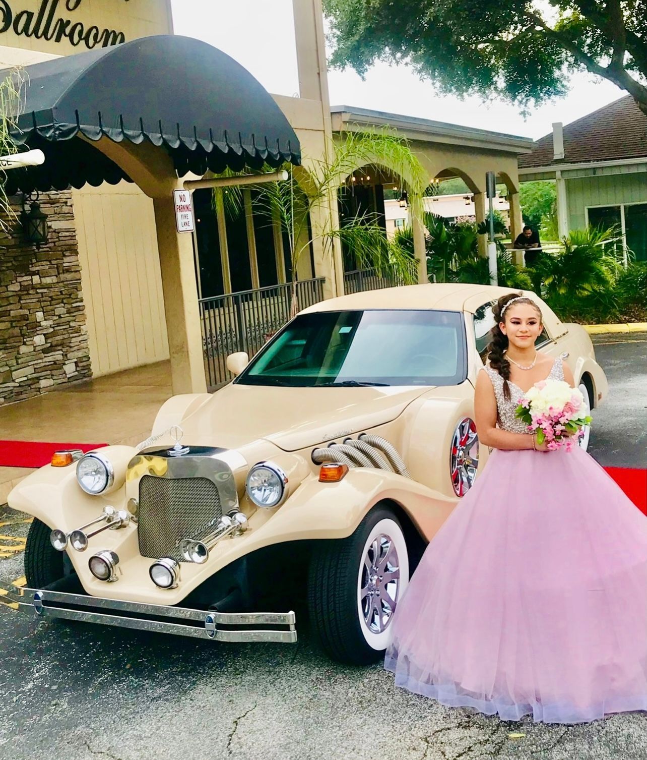 A person in a formal, light pink ballgown holds a bouquet next to a cream-colored vintage-style car in front of a building.