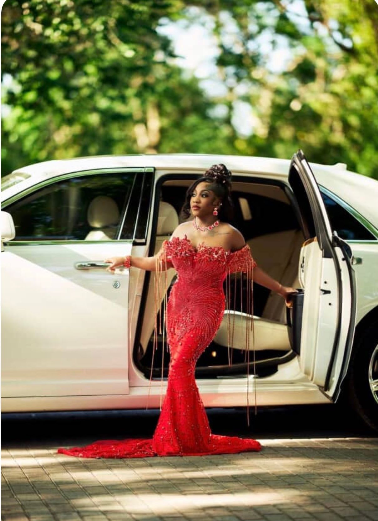 A person in a red, off-the-shoulder sequined gown with fringe details standing beside an open white luxury car door.