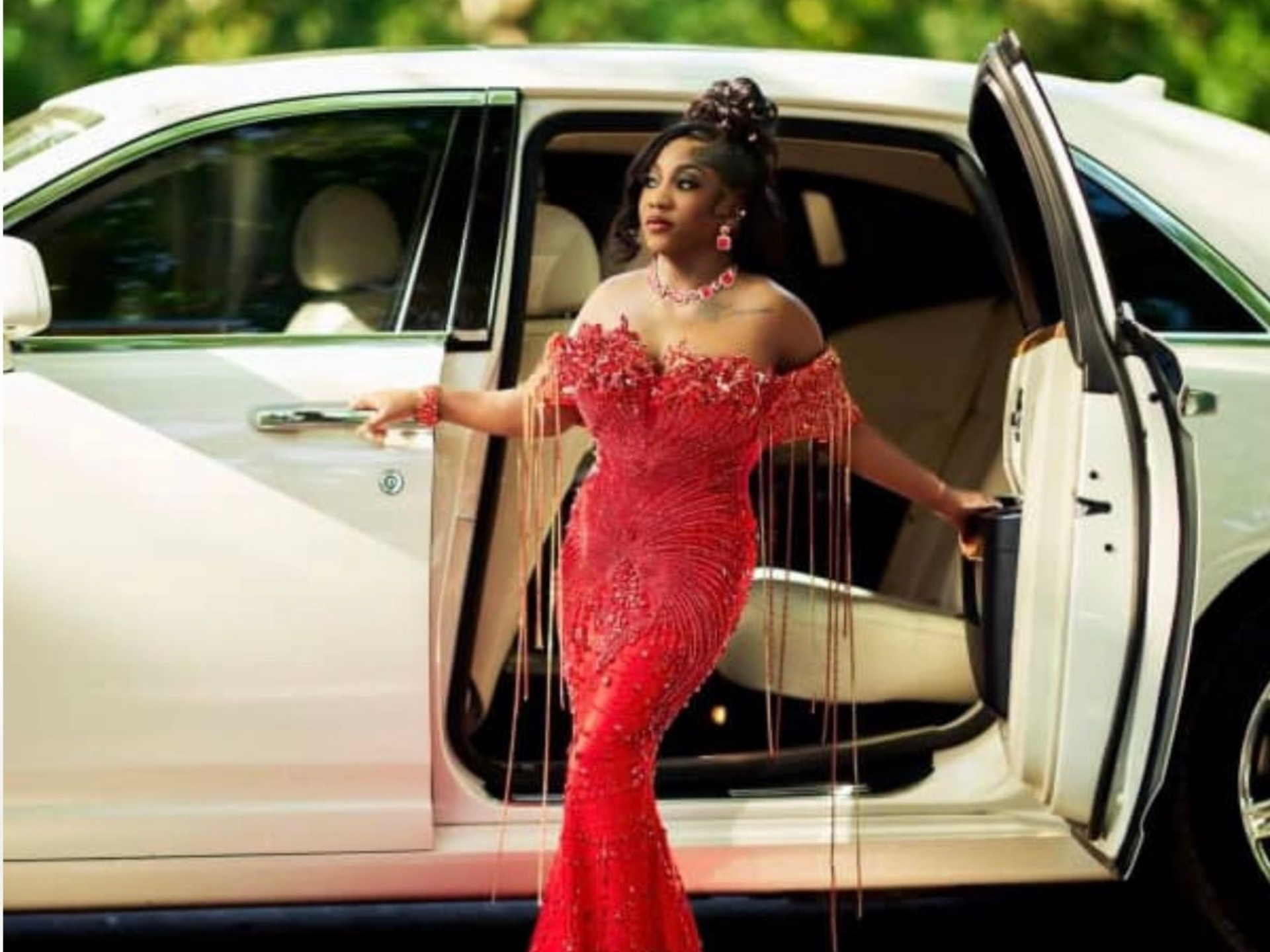 A person in a red, off-the-shoulder beaded gown with fringe details steps out of a white luxury car.