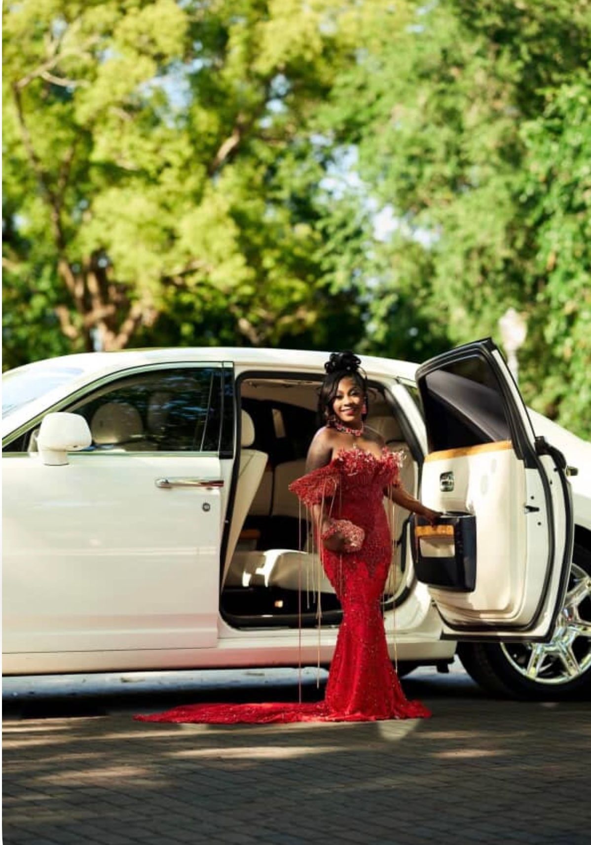 A person in a red off-the-shoulder gown standing beside an open white limousine on a paved surface with trees behind.