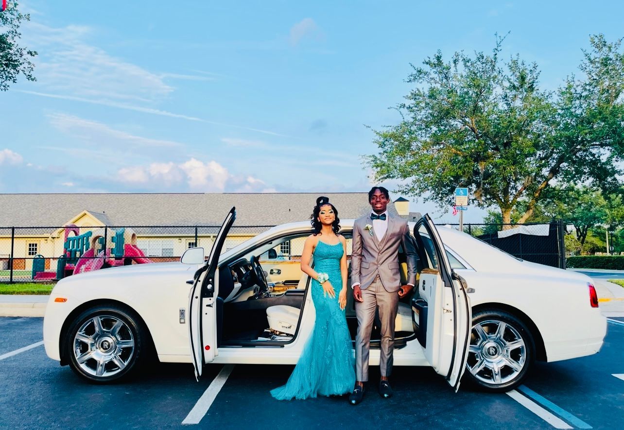 A couple dressed for a formal event stands beside a white luxury car with its doors open in an outdoor parking lot.