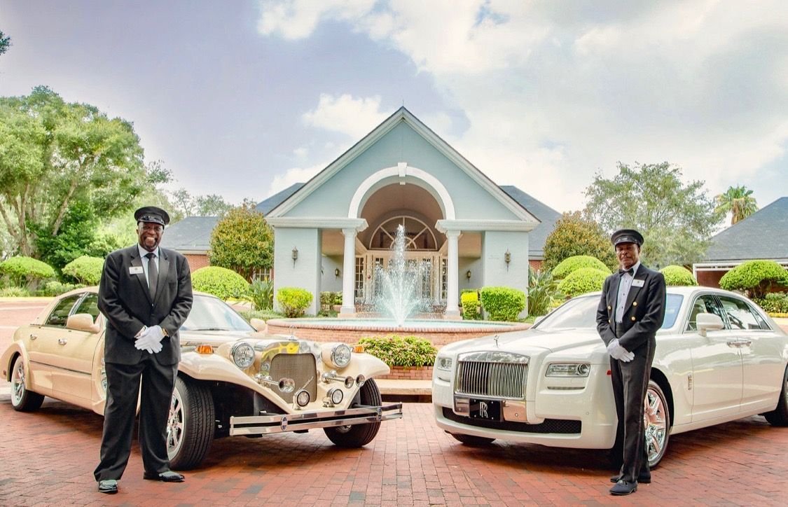 Two uniformed chauffeurs stand in front of luxury cars parked before a building with a courtyard fountain.