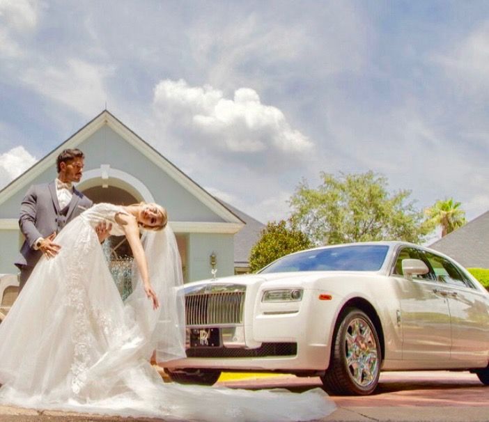 A bride in a white wedding gown performs a dip with her partner in front of a white luxury car outside a building.