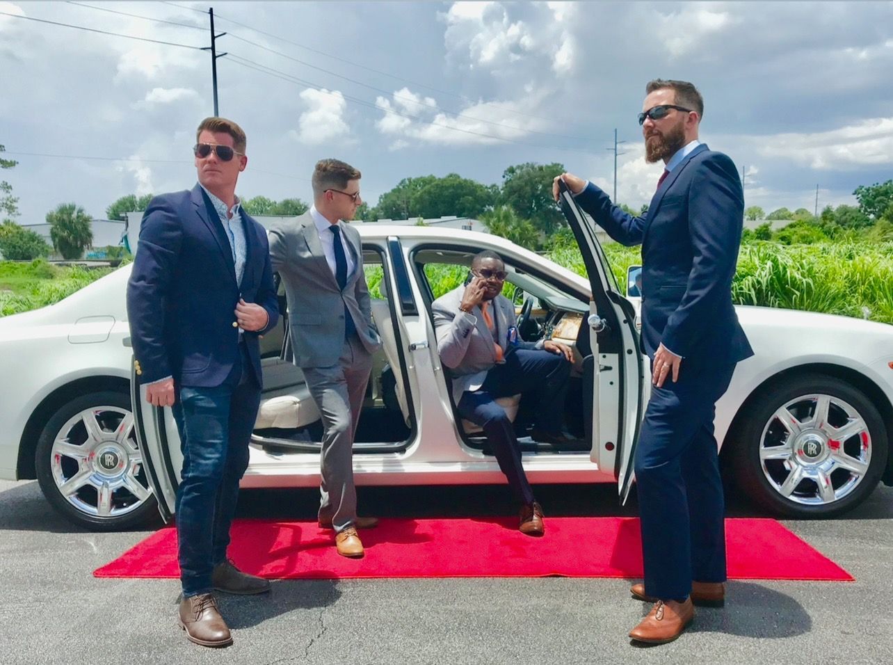 Four men in suits stand near a white luxury car on a red carpet outdoors on a sunny day.
