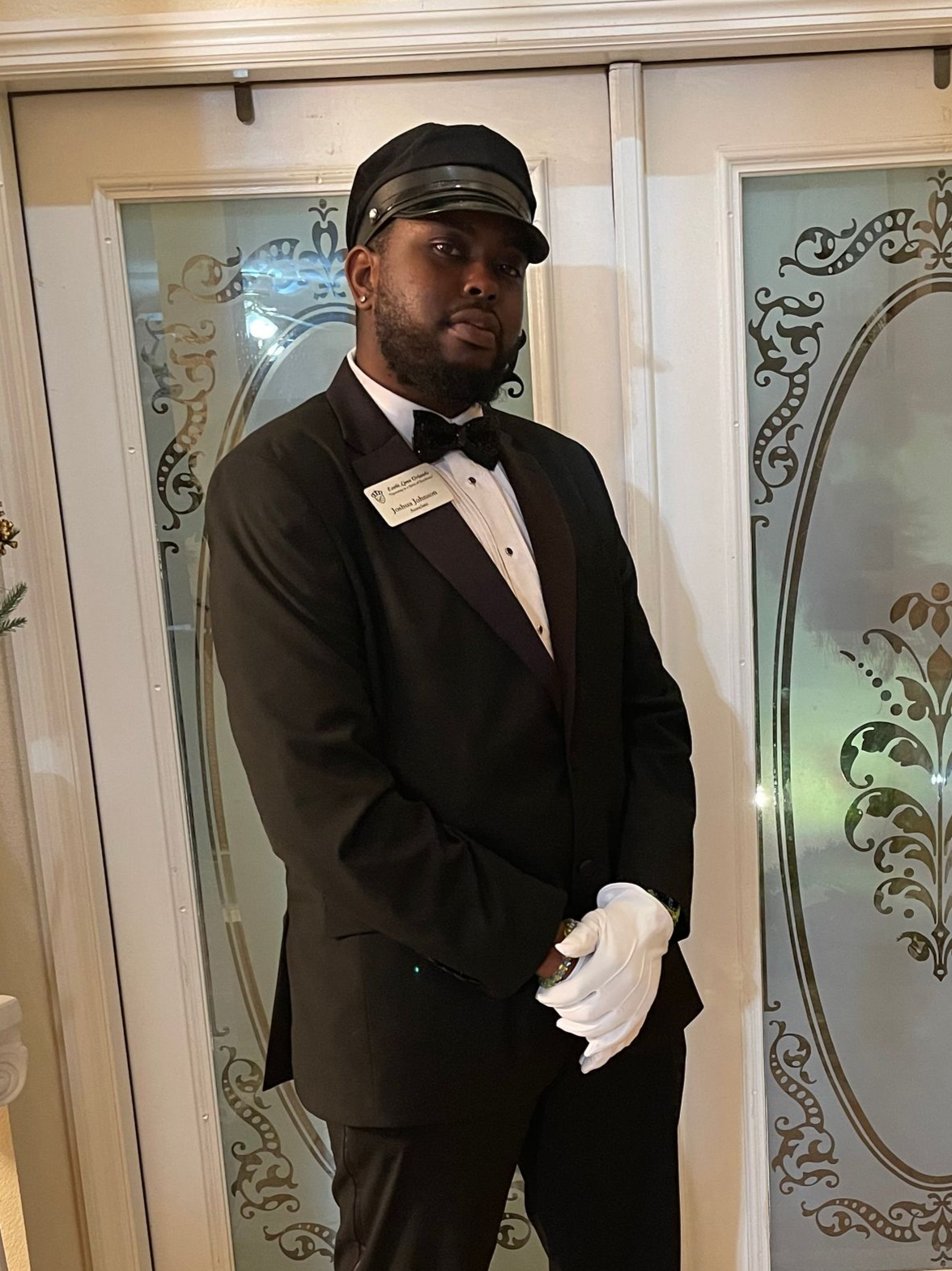 A person wearing a tuxedo, bow tie, white gloves, and a black cap stands with hands clasped in front of double doors.
