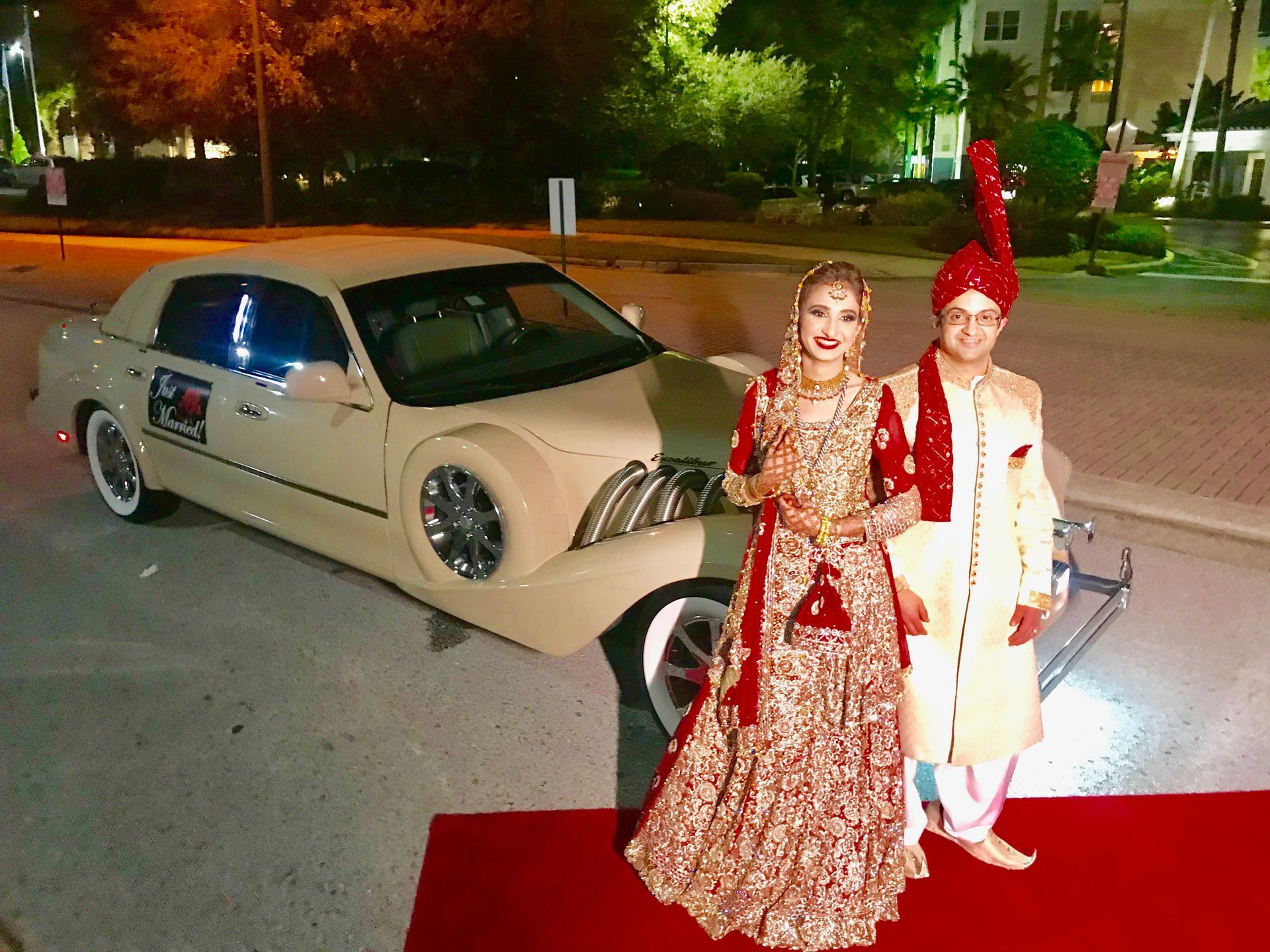 A couple in wedding attire poses next to a decorated beige limousine on a red carpet at night.
