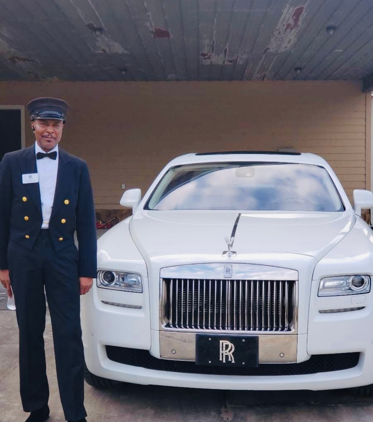 A chauffeur in a dark suit and hat stands beside a white Rolls-Royce parked in front of a building.