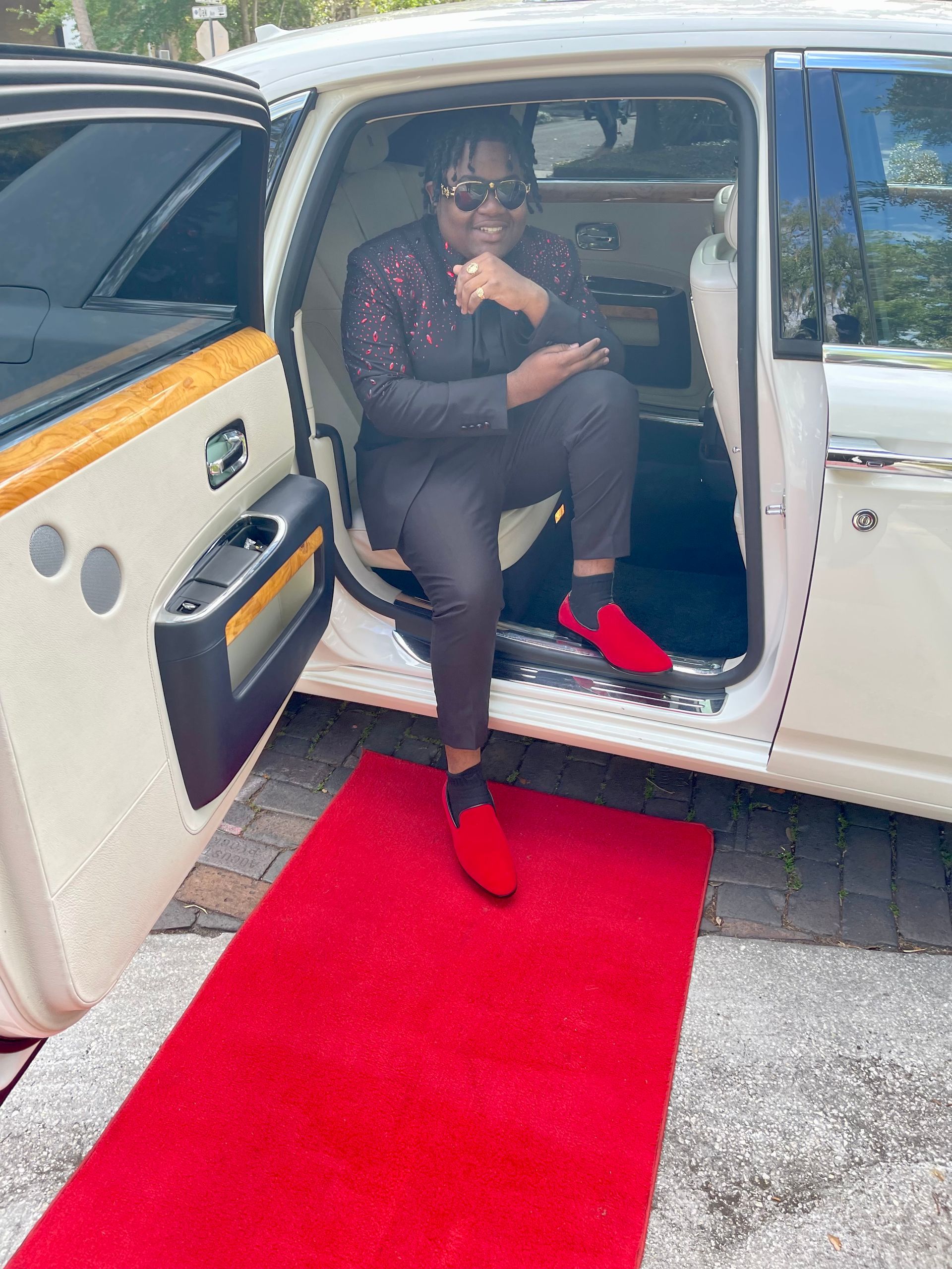 A person wearing a suit and red shoes sits in a white luxury car door opening, with a red carpet rolled out in front.