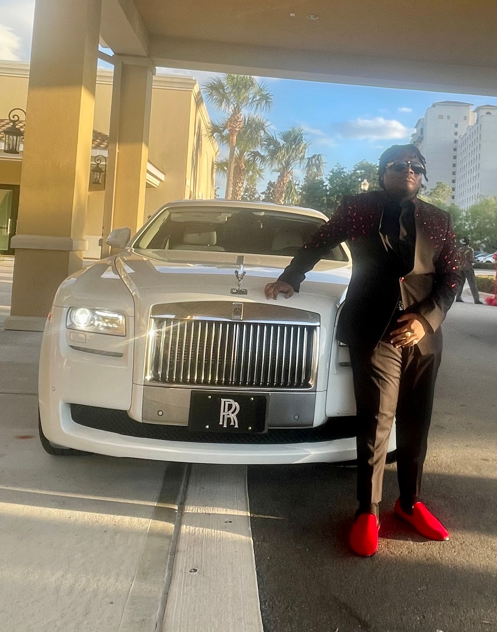 A person in a dark suit and red sneakers stands beside a white Rolls-Royce parked under an outdoor covered walkway.
