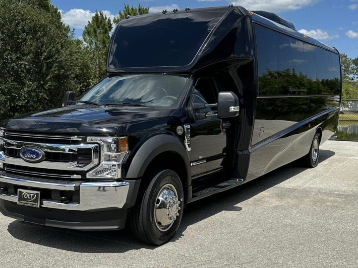 A black Ford F-series shuttle bus parked on a paved surface against a blue sky with trees in the background.