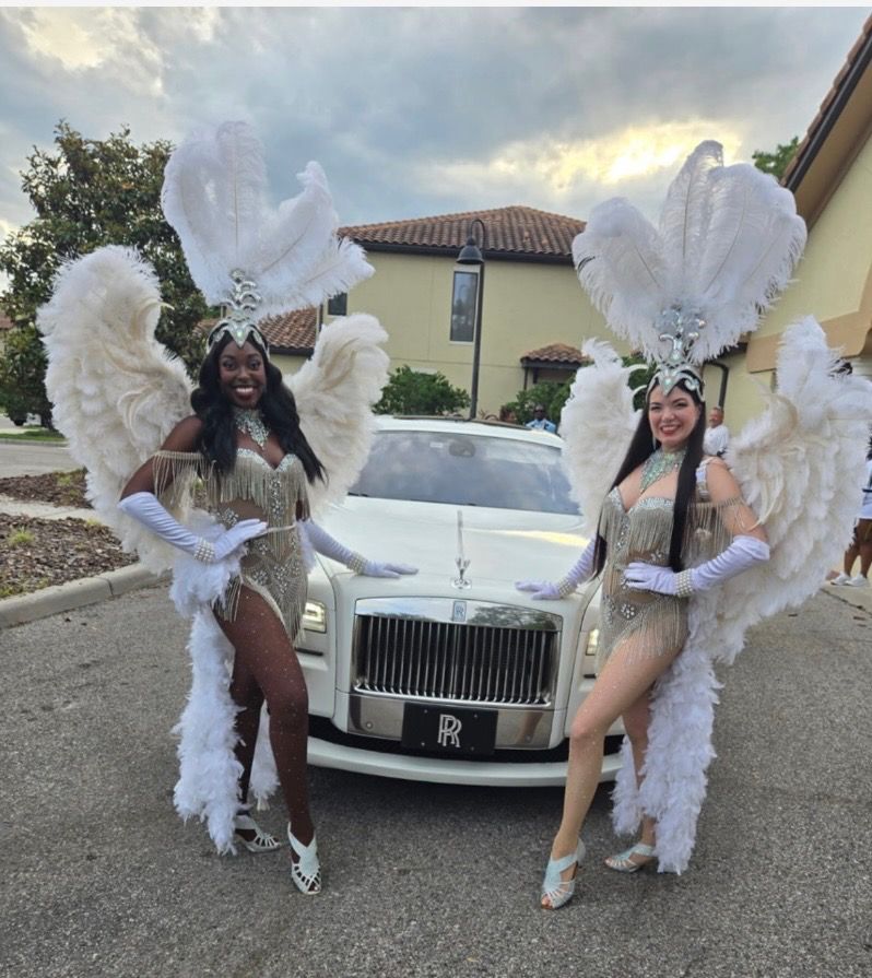 Two performers in ornate feather wings and white costumes pose with a white Rolls-Royce in a parking lot.