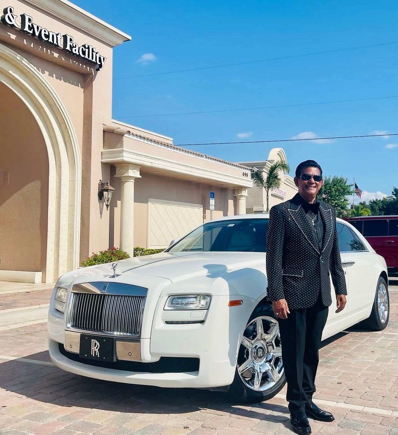 A person in a patterned blazer stands smiling next to a white luxury car parked in front of a building.