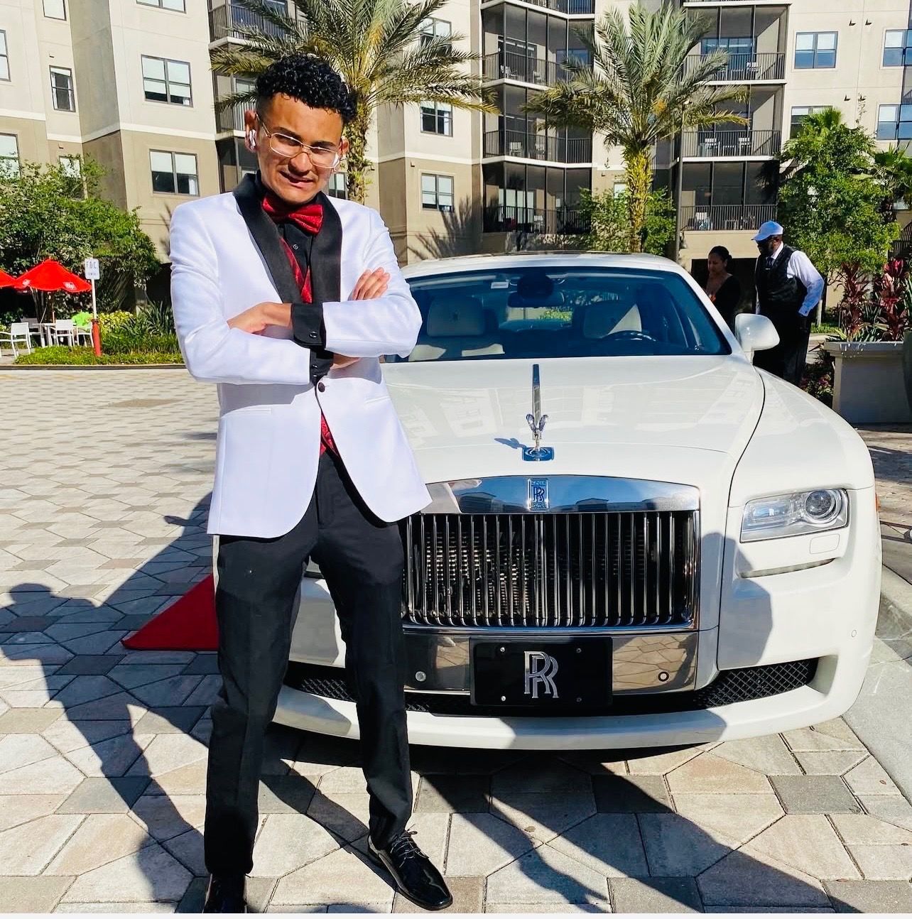 A person in a white suit with a red bowtie and shirt stands with arms crossed in front of a white Rolls-Royce.