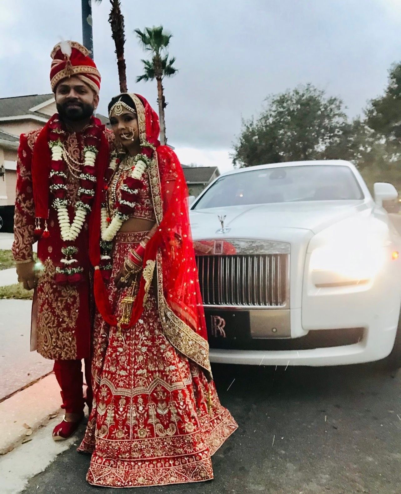 A couple in traditional red wedding attire stands next to a white Rolls-Royce parked on a residential street.