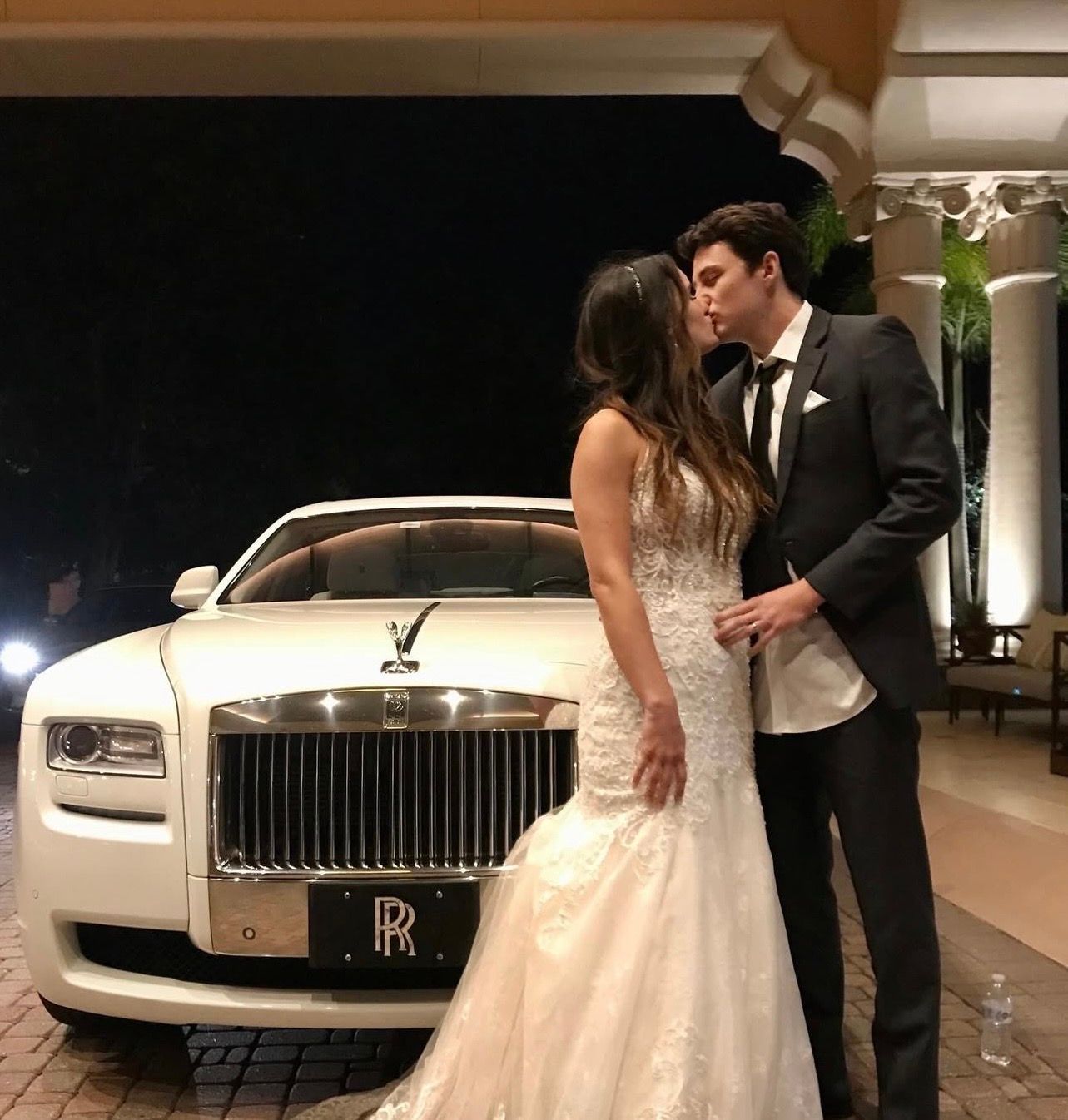 A bride and groom in wedding attire kiss in front of a white Rolls-Royce at night.