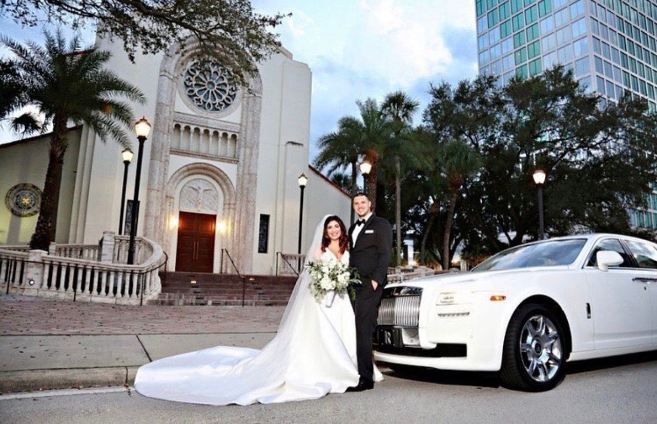 A couple in wedding attire poses in front of a white luxury car outside a stone church at twilight.