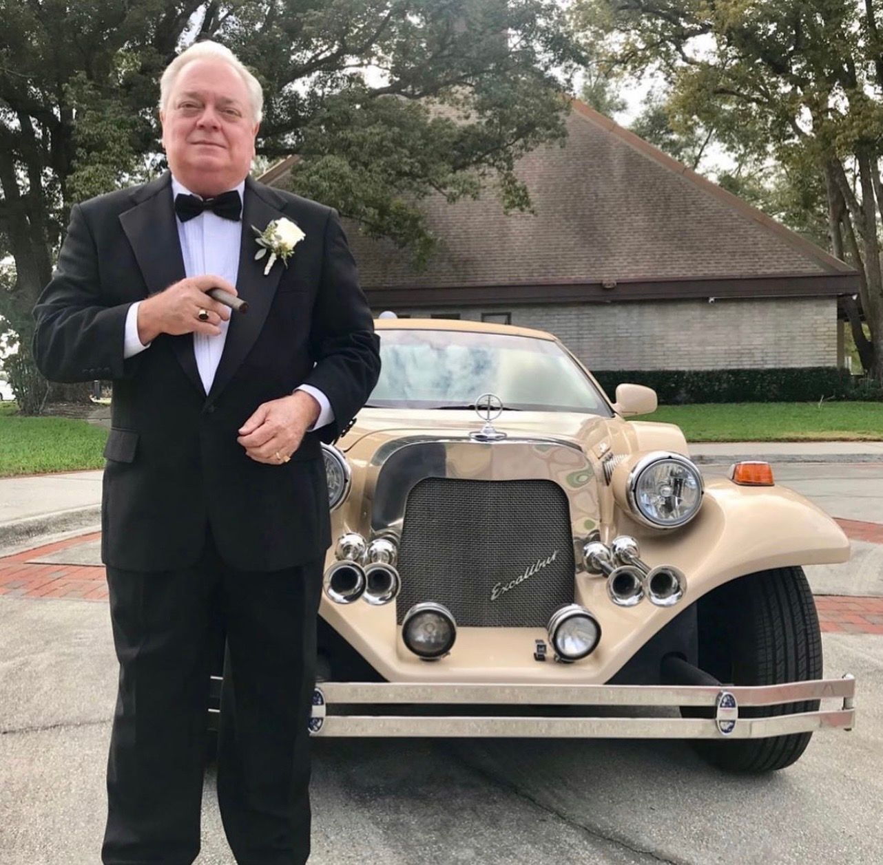 A person in a black tuxedo with a boutonnière holds a cigar in front of a beige vintage-style convertible car.