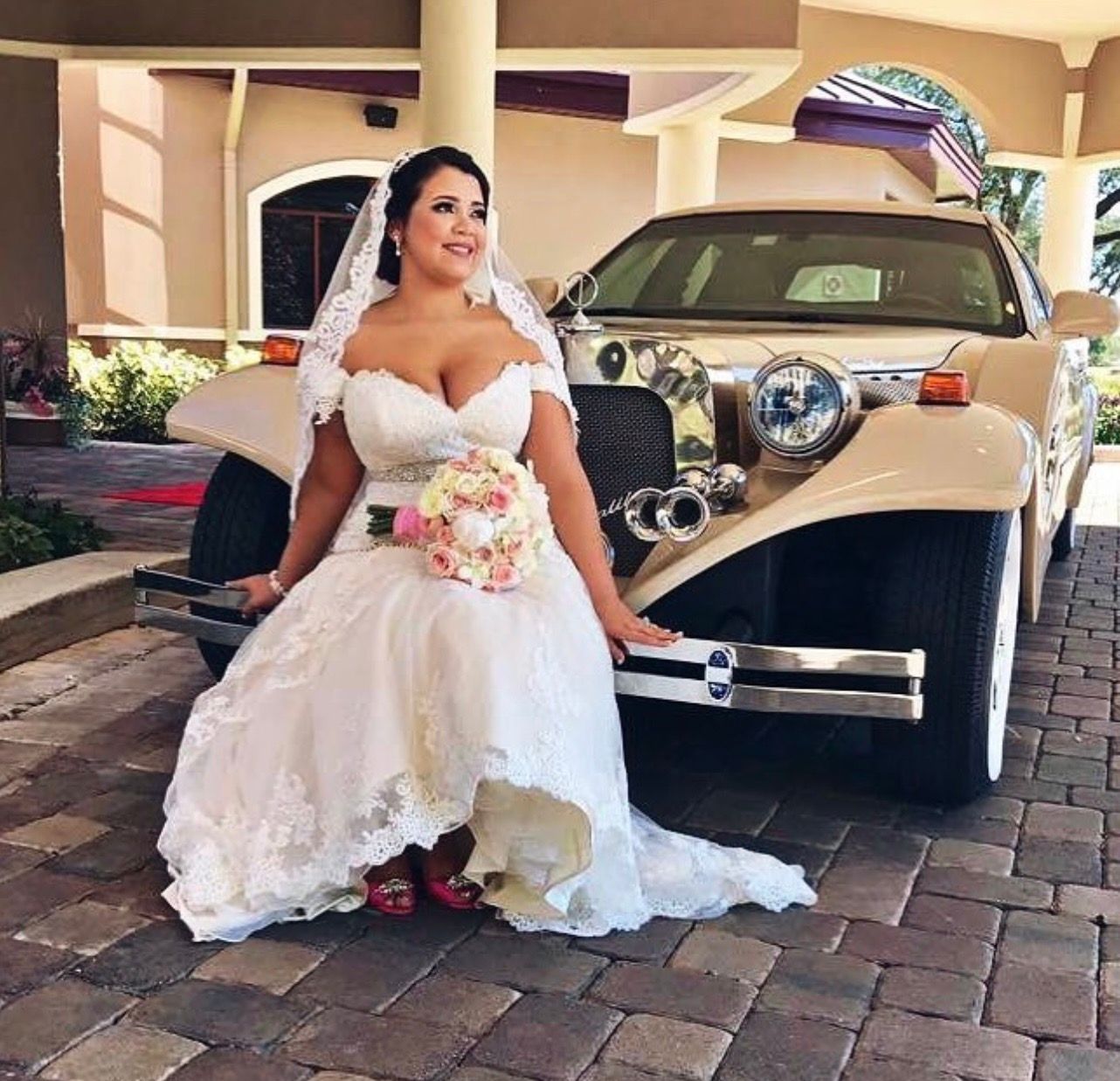 A bride in a white wedding gown and veil sits on the front bumper of a classic beige vintage car.